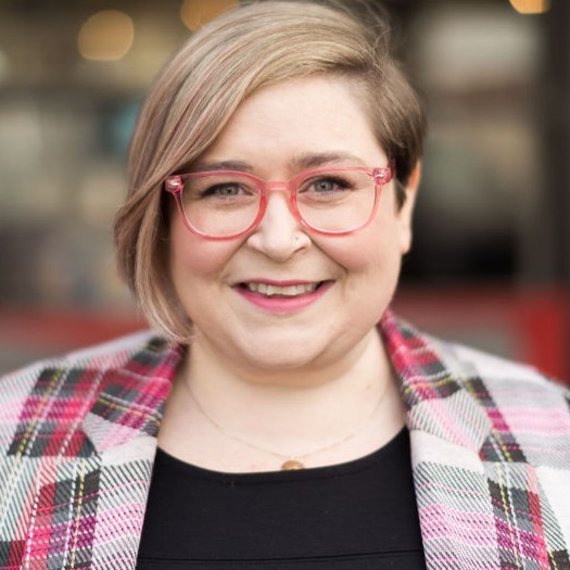Headshot of Kate. A woman with light freckly skin, a blonde bob with a hint of pink, and pink-framed glasses.
